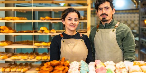Two bakers standing behind a display of pastries and bread in a bakery.Having just obtaining Equipment funding for their business. 