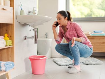 Woman on phone checking leaking sink with bucket below.