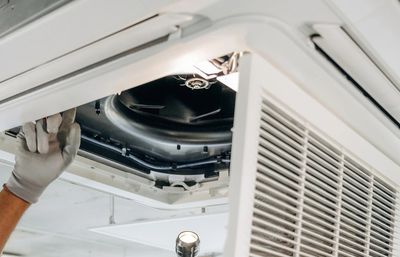Technician inspecting and repairing a ceiling air conditioning unit with a flashlight.