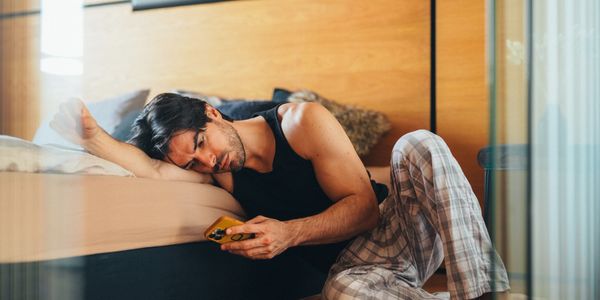 A man in pajamas lying beside a bed, looking at his phone with a tired expression.