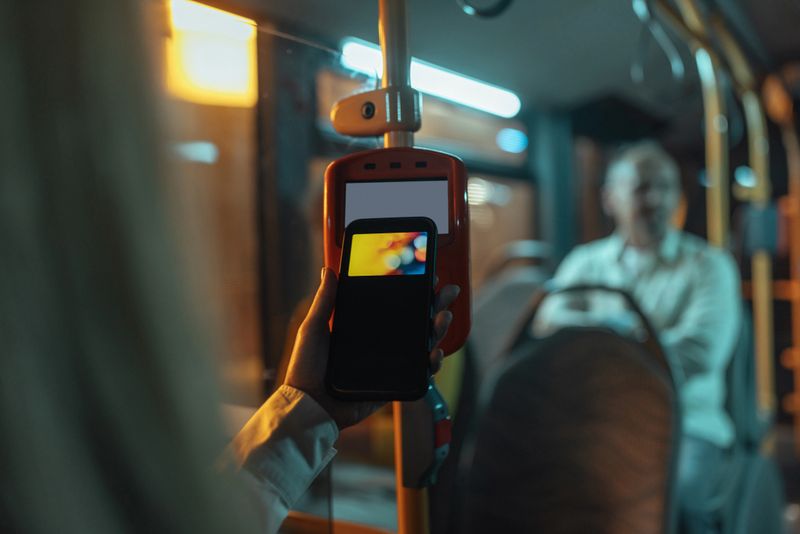 Close up of a person paying the bus ticket via a smarthpone terminal at the front doors with the other passengers visible in the blurred background.