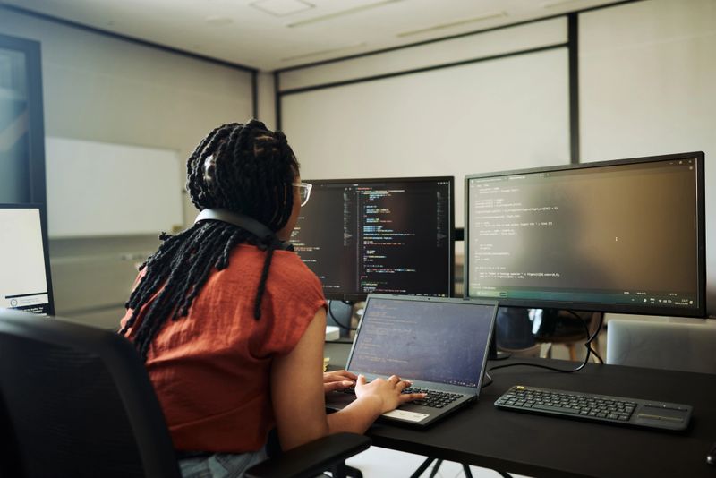 Young woman programmer focused on her work, coding on dual monitors in a modern office environment