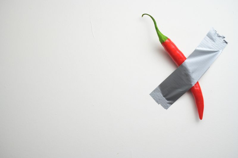 A vibrant red chili pepper secured to a white wall with duct tape, symbolizing bold creativity and conceptual art humor.