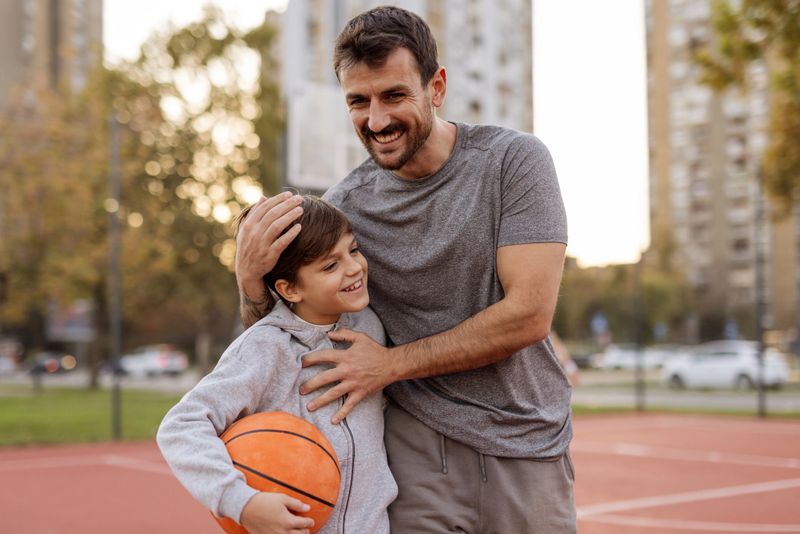 After a fun and energetic outdoor basketball practice, a proud father wraps his arms around his little son in a heartfelt hug. The son, smiling brightly, leans into the embrace, clearly enjoying the moment of connection. Their faces show pride and happiness, capturing a joyful moment of accomplishment and love after a successful practice.
