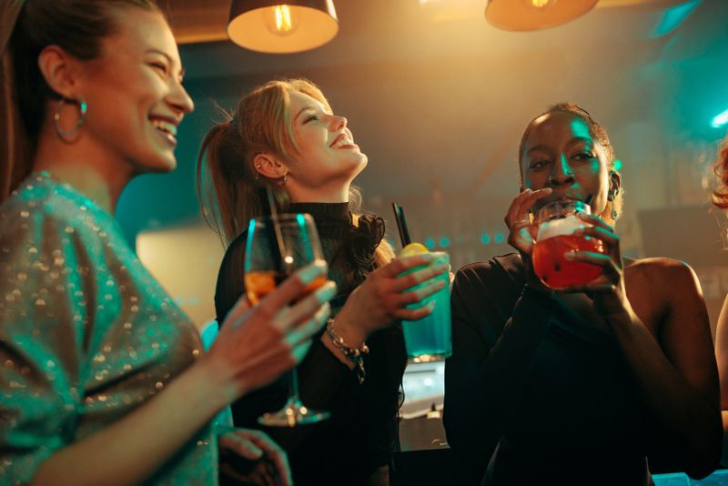 A multiracial group of girls smiling and sipping their cocktail drinks, standing by the bar in a crowded nightclub.