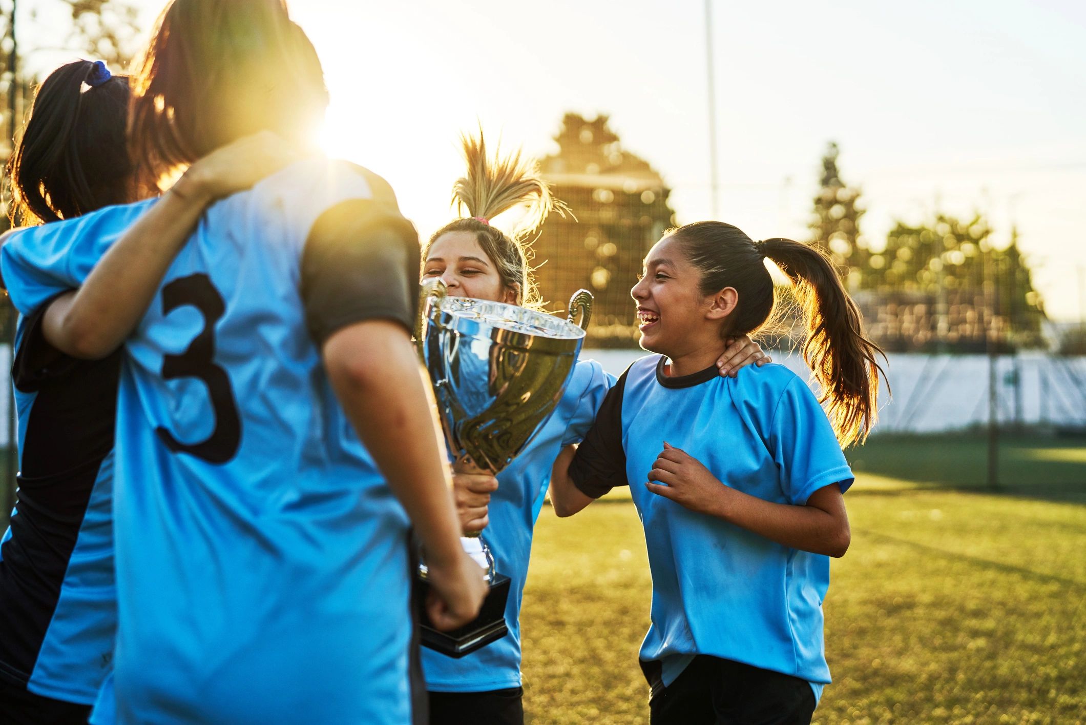 Girls soccer team celebrating with a trophy at sunset.