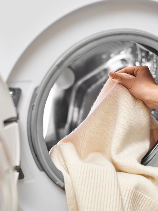 Hand placing a beige sweater into a front-loading washing machine.