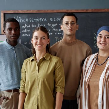 Diverse group of four young adults smiling in front of a chalkboard in a classroom.