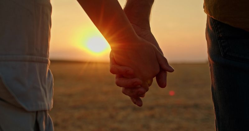Close-up of two hands holding each other at sunset in an open field, symbolizing connection, love, and support. The golden light of the setting sun creates a warm and intimate atmosphere. High quality photo