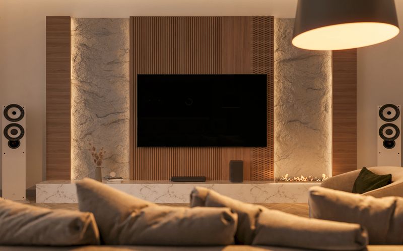 The image presents a contemporary living room in warm, neutral tones. A large television mounted on a wood and stone wall serves as the centerpiece, flanked by high-quality speakers to enhance the audio-visual experience. Thoughtful lighting creates a cozy ambiance. The minimalist furniture highlights natural materials, contributing to an inviting and spacious atmosphere. 3D render