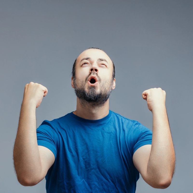 Excited man with a beard and blue t-shirt is raising his fists in the air, celebrating a victory with an expression of pure joy and excitement