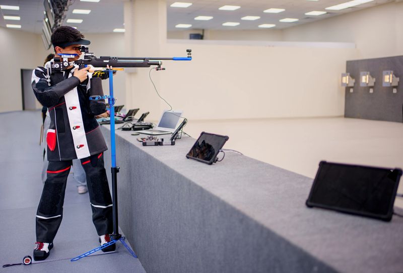 A focused athlete practicing precision shooting in an indoor shooting range. The athlete is equipped with specialized gear, including a custom rifle, shooting jacket, and protective eyewear. The setting includes advanced digital target monitoring systems and training equipment, reflecting a professional training environment. Ideal for themes of sports, dedication, and high-performance training.