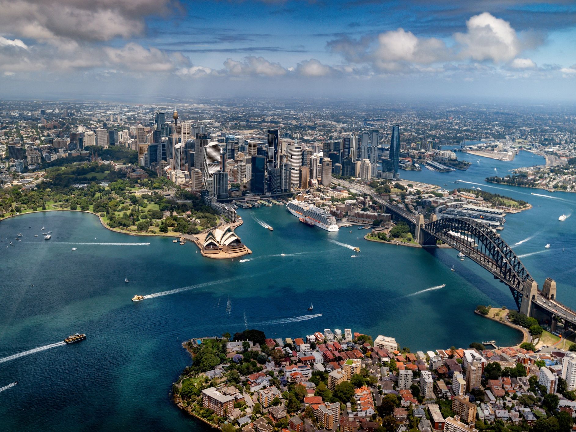 Aerial view of Sydney's iconic Opera House, Harbour Bridge, and city skyline under a partly cloudy sky.