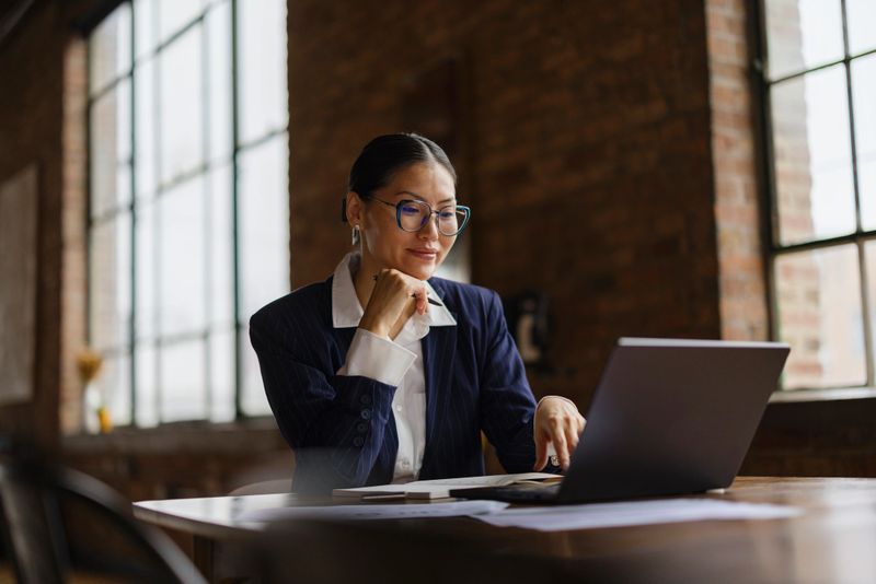 A woman in smart casual attire focuses on her laptop in a modern office. The bright, urban environment suggests productivity and professionalism. Glasses add a scholarly touch to the scene.