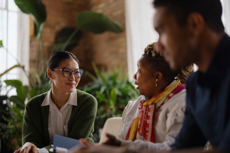 A diverse group of colleagues engage in a conversation in a bright, green office setting. They are dressed in smart casual attire, creating a welcoming and collaborative work environment.