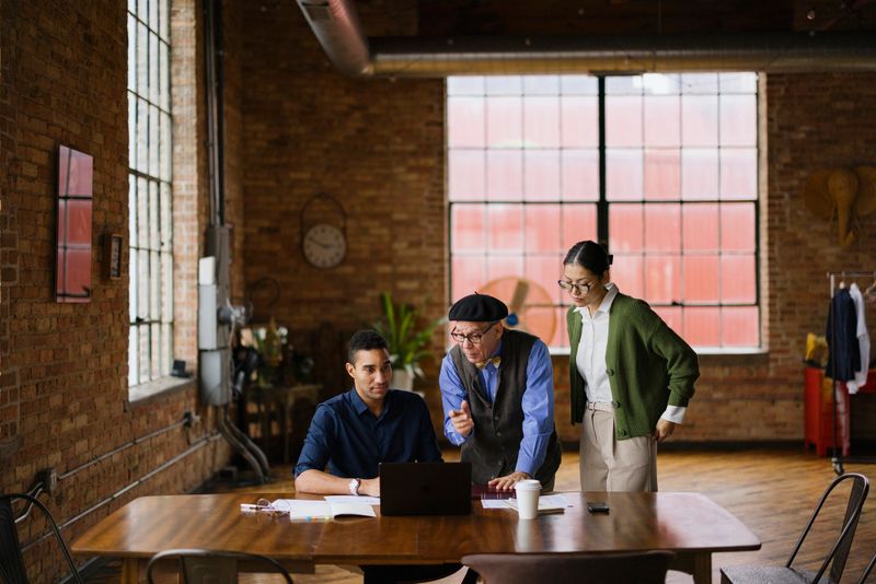 A diverse group of three professionals engaged in a discussion around a laptop in a stylish office. They appear focused and collaborative, working on a project in a modern industrial setting.