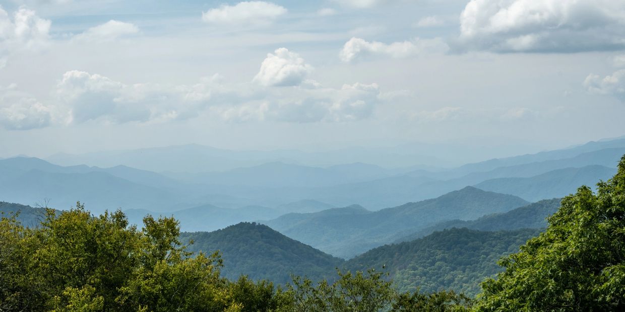 Layered blue mountains under a cloudy sky with green trees in the foreground.