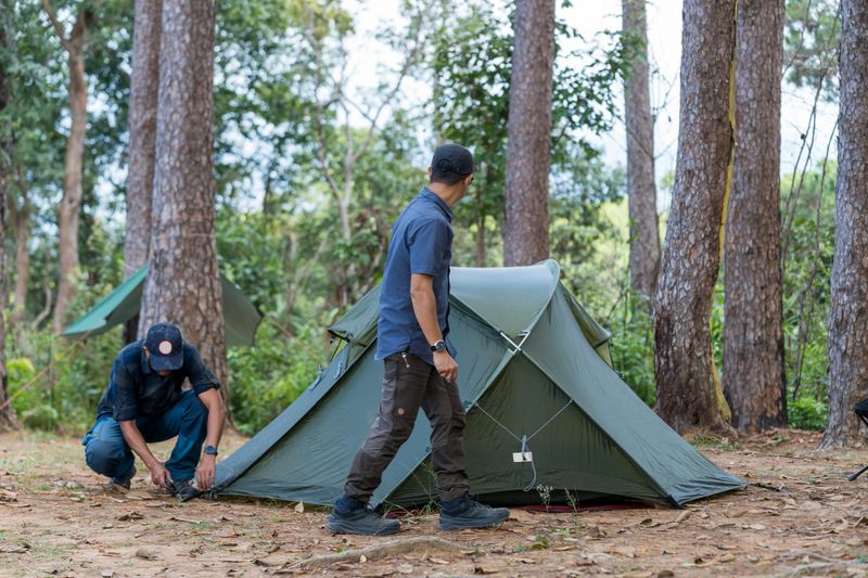 Man wearing a hat, setting up his tent in the forest during sunset, preparing for camping adventure in the wilderness. Camping, tourism and travel concept, man setting up tent outdoors