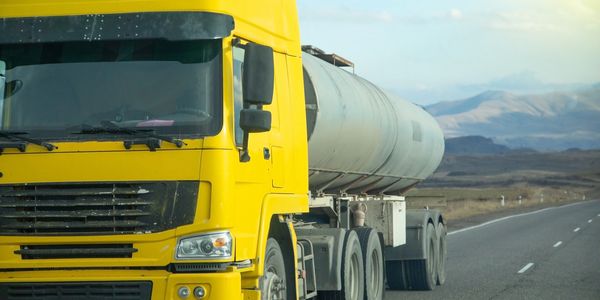 Yellow tanker truck driving on an open highway with mountains in the background.