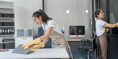 Two women cleaning an office space, one wiping a desk and the other a glass wall.