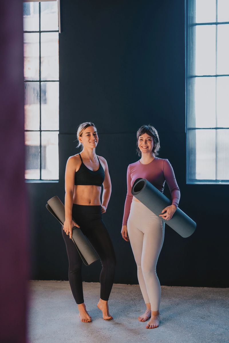 Two women stand confidently holding yoga mats in a sunlit fitness studio, embodying health and well-being. Their smiles reflect a commitment to an active lifestyle and mindfulness.
