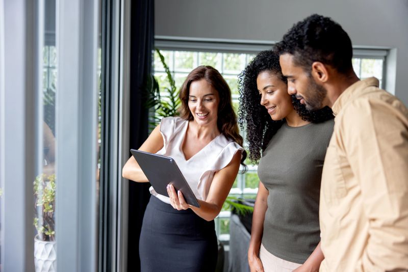 Happy real estate agent showing a couple some pictures using a digital tablet while looking at a house for sale