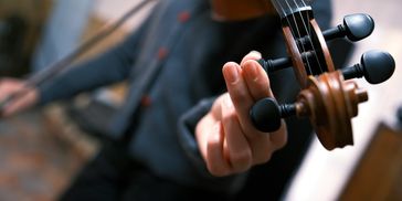Close-up of a person tuning a violin's peg with a bow in the background.
