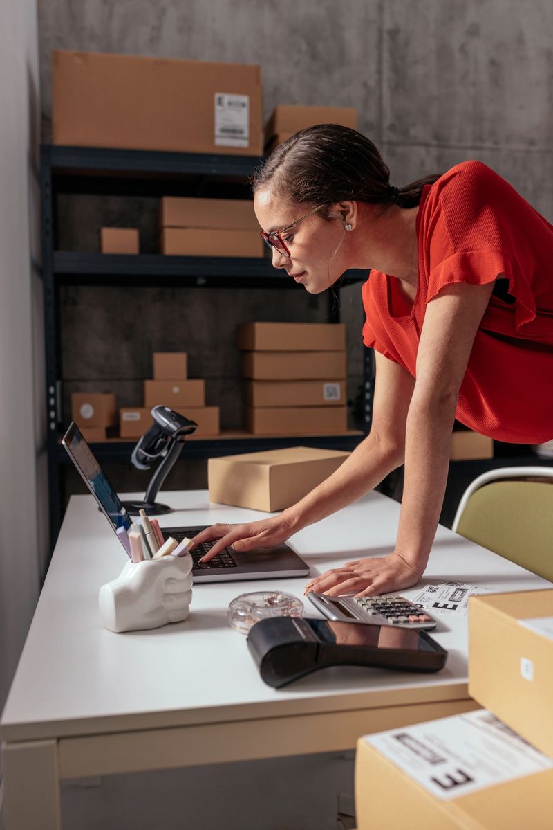 A woman in a red shirt is engaging with logistics in a warehouse, organizing parcels and using a laptop for tracking