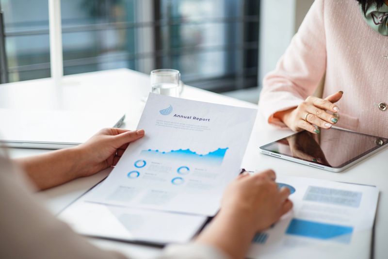 Two businesswomen analyze an annual report during a meeting, highlighting teamwork and strategy. The use of a digital tablet emphasizes the modern, technology-driven office environment.