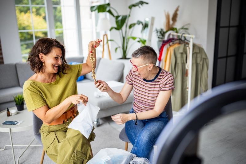 The women share a vibrant moment as one displays a beautifully designed necklace, highlighting themes of friendship, fashion, and creativity in a stylish environment.