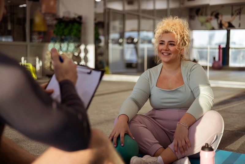 A young, plus-sized woman is engaged in a conversation with a male fitness instructor at the gym. The instructor, dressed in athletic gear, is offering guidance and sharing fitness tips with her. The woman listens attentively, looking motivated and focused on her fitness journey. The gym is well-lit and equipped with various workout machines in the background. This image captures a supportive coaching moment, highlighting the importance of professional guidance and encouragement in helping individuals, especially those aiming for weight loss and improved health, achieve their fitness goals