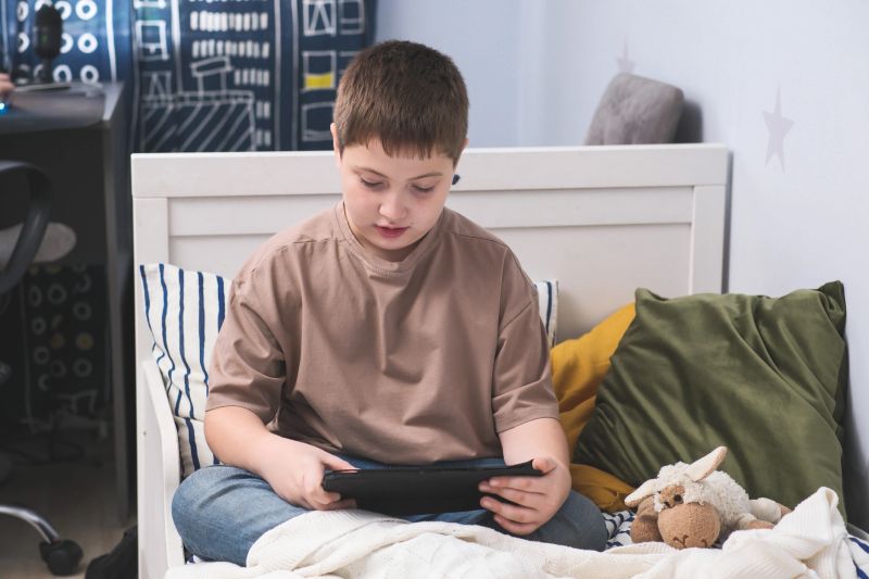 A young boy sits on his bed, focused on his tablet, plays videogame, surrounded by soft pillows and a plush toy, creating coziness