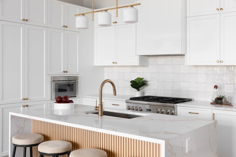 A kitchen detail with white cabinets and square tile backsplash, a waterfall marble countertop and wood slat panel front, and a gold light fixture, hardware, and faucet. No brands or labels.