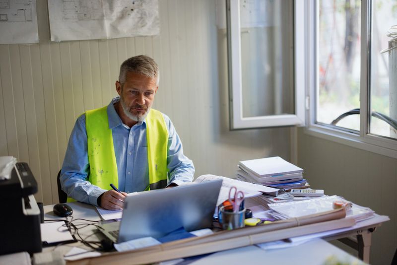 A construction manager in a safety vest studies building plans at his desk. Natural light fills the room, highlighting the papers and equipment around him. He is focused on project details.