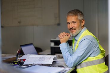 Smiling engineer in a yellow safety vest at a desk with blueprints and a laptop.