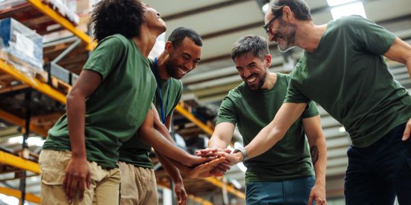 Group of workers in green shirts stacking hands in a warehouse.