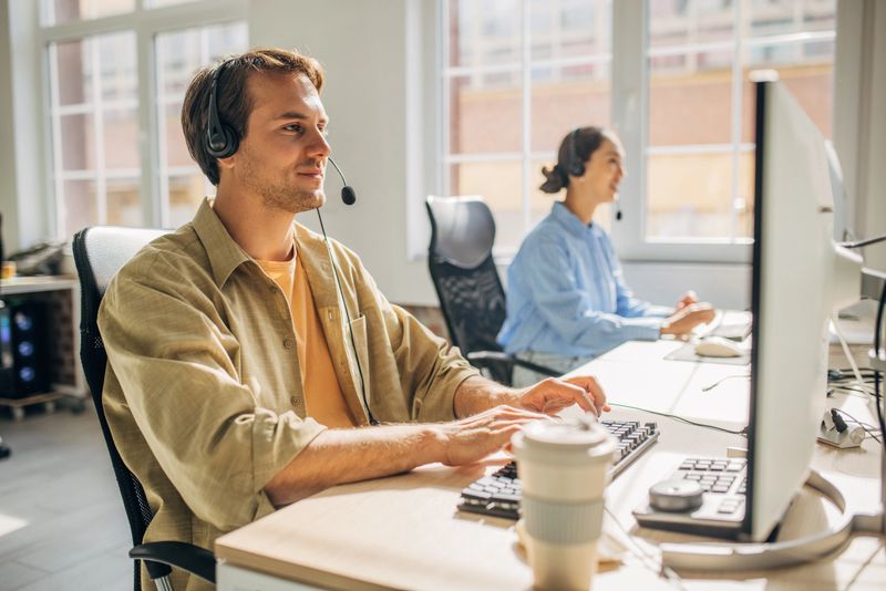 In a well-lit office, a male customer service agent speaks animatedly while working at a computer, providing an engaging interaction with clients in a modern workspace.