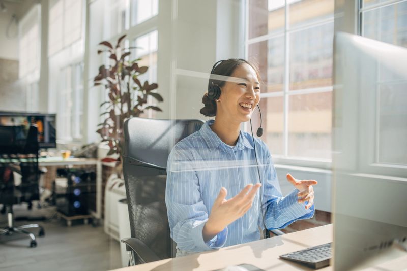 A happy female call center agent is conducting a video call with her client. She smiles warmly while discussing in her bright office space filled with natural light.