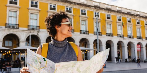 A happy tourist with sunglasses and yellow backpack reads a map in a sunny plaza.