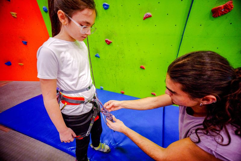 An experienced climber helps a young girl adjust her climbing harness in a vibrant indoor climbing gym. The adult is seated on a padded mat while providing guidance, ensuring the harness is secure. The girl, standing confidently in climbing shoes and gear, prepares for her climbing experience. The colorful climbing wall and ropes in the background highlight the engaging and educational environment. This scene emphasizes safety, mentorship, and the introduction of climbing to children in a supportive setting