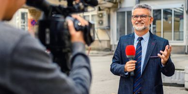 Male reporter with glasses holding a microphone during an outdoor video shoot.