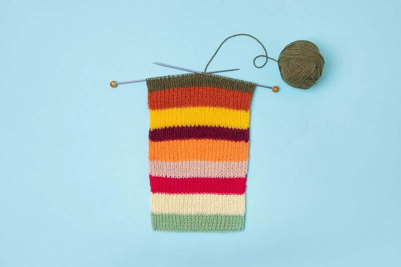 Green knitted wool on turquoise background with knitting needles for knitting warm clothes and hobbies needlework. Knitting striped bright scarf made of red, yellow, green, orange and purple threads.