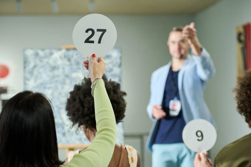 Rear view of young brunette woman raising hand holding paddle with number twenty seven while taking part in bidding