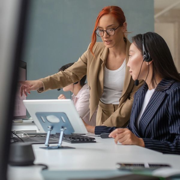 A woman in a tan blazer instructs a colleague wearing a headset in an office.
