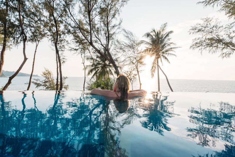 Woman leaning on the edge of an infinity pool surrounded by tropical trees, gazing at the ocean as the sun sets, creating a serene and tranquil ambiance.