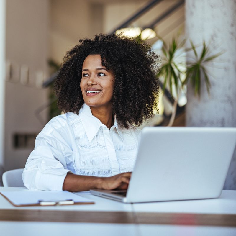 A cheerful young professional with curly hair sits at her desk in a modern office, working on her laptop and smiling, indicating a moment of happiness and successful productivity.