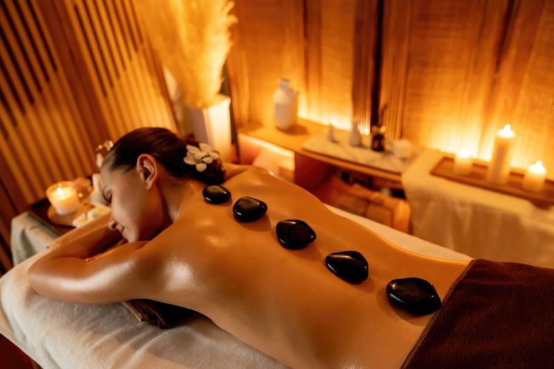 Hot stone massage at spa salon in luxury resort with warm candle light, blissful woman customer enjoying spa basalt stone massage glide over body with soothing warmth. Quiescent