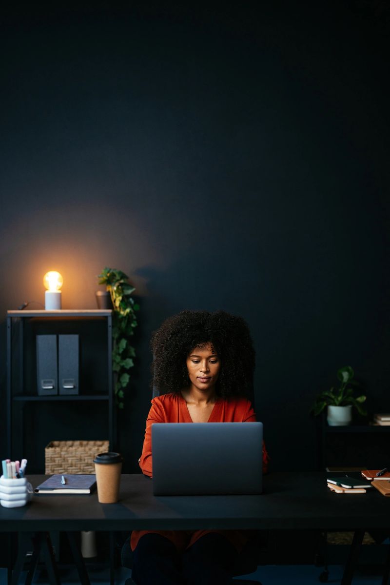 Young woman concentrating on her work at a laptop in a dimly lit, modern office, embodying focus and ambition in her professional environment