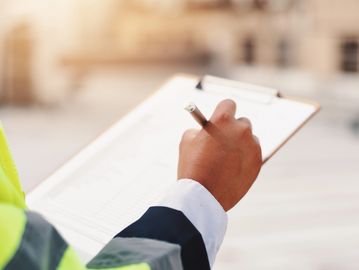 Person in safety jacket writing on a clipboard outdoors.