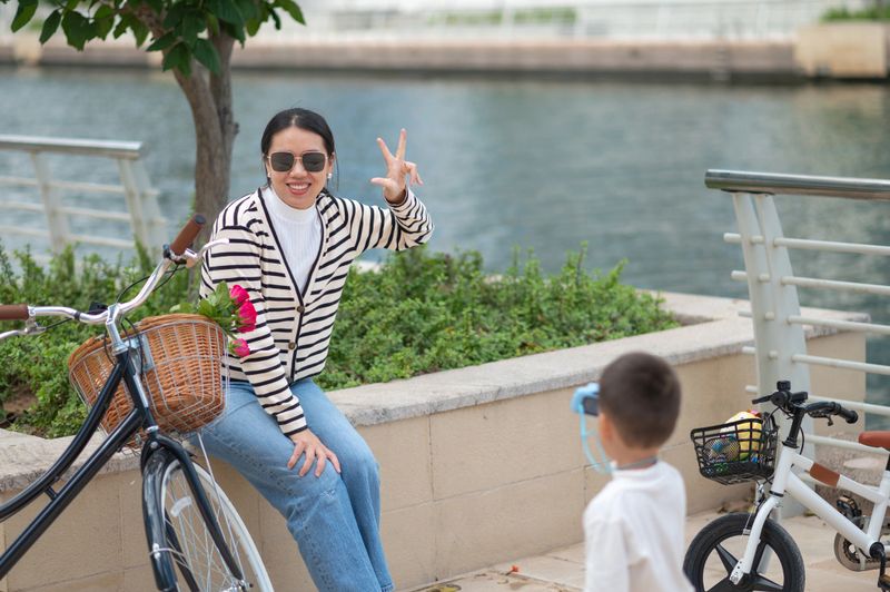 3-year-old boy capturing a photo of his mother with a small camera during a family bicycle outing by a scenic canal. The moment reflects family bonding, curiosity, and childhood creativity by the water